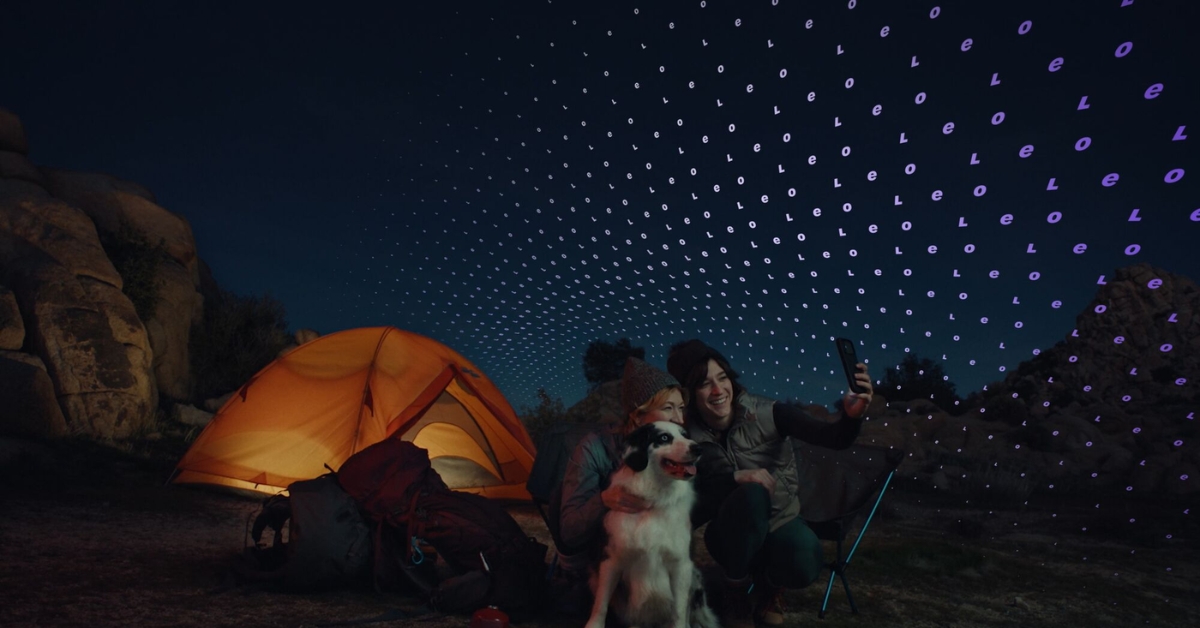 Two friends camping at night pose for a selfie with their dog beside an orange tent, while a grid of purple light dots fills the sky.