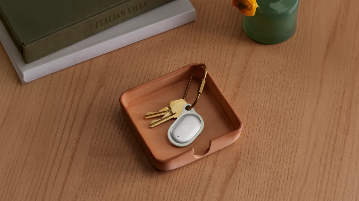 Keys and a gray key fob resting on a small copper tray on a wooden desk, with books and a green vase nearby.