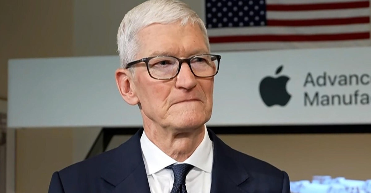 Older man in a navy suit and glasses at a manufacturing event, with an Apple logo and 'Advanced Manufacturing' sign in the background.]