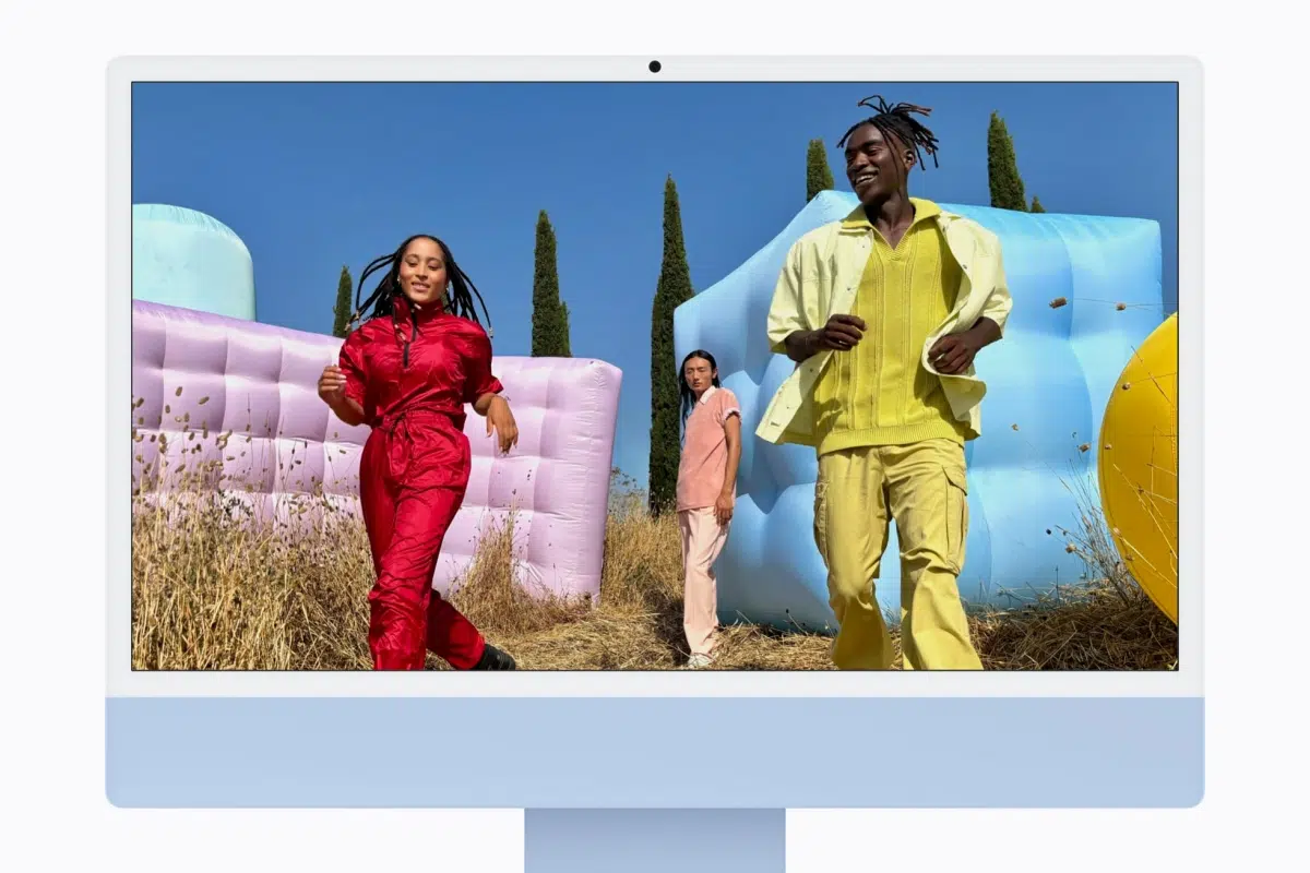 Apple iMac M6 launched: Specs, price, and release date Three people in bright outfits walk past pastel inflatable structures outdoors on a sunny day, with dry grass in the foreground and blue sky behind them.