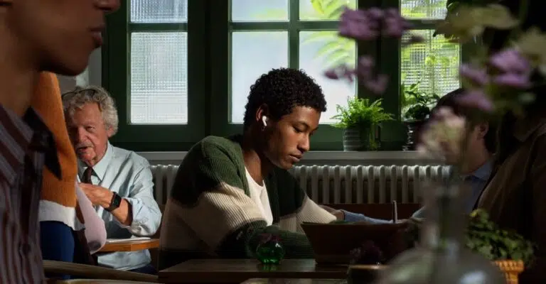 A man in a green cardigan wears AirPods 5 while reading at a café table in soft window light