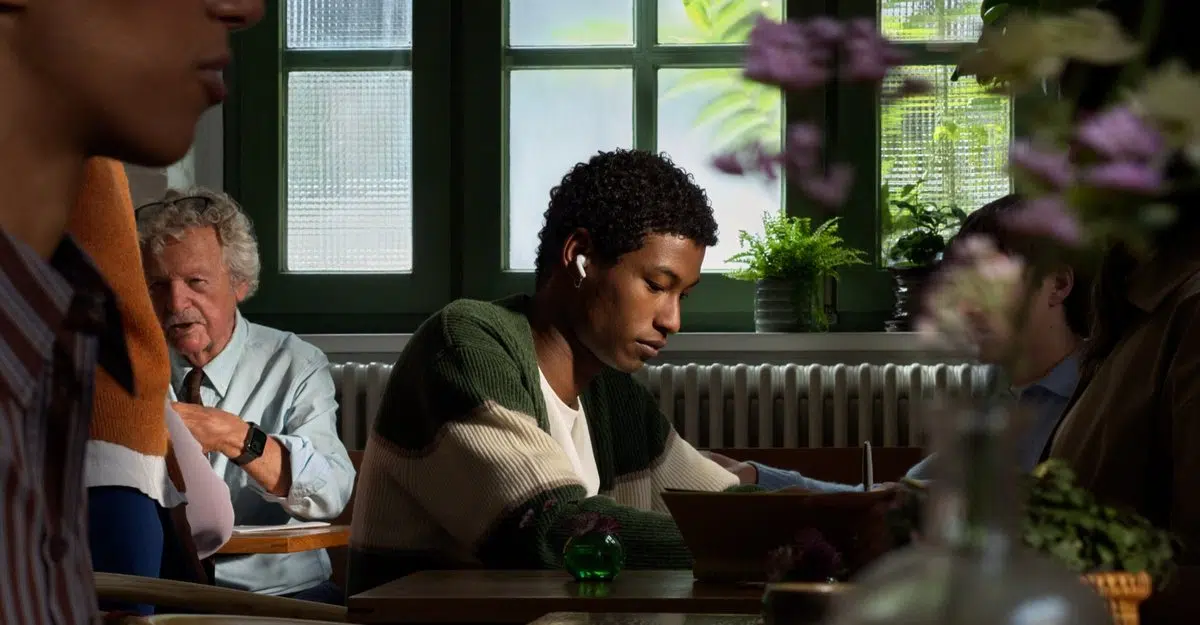 A man in a green cardigan wears AirPods 5 while reading at a café table in soft window light