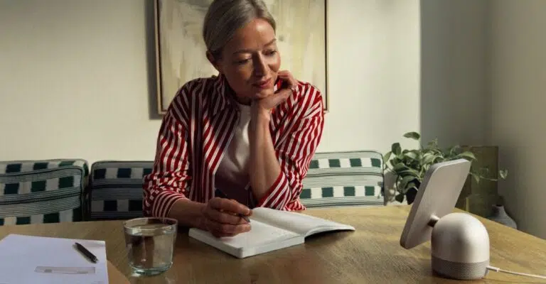 Woman writing in a notebook at a wooden dining table beside an Apple HomeView tabletop display