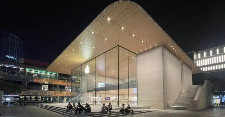 Apple Store Sanlitun in Beijing lit up at night, with its wooden canopy over a glass front and people sitting on the steps.