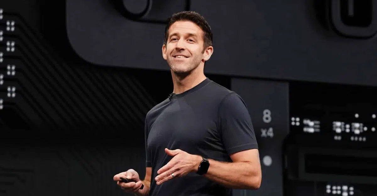 John Ternus smiling and gesturing on a dark stage in front of a circuit-style backdrop
